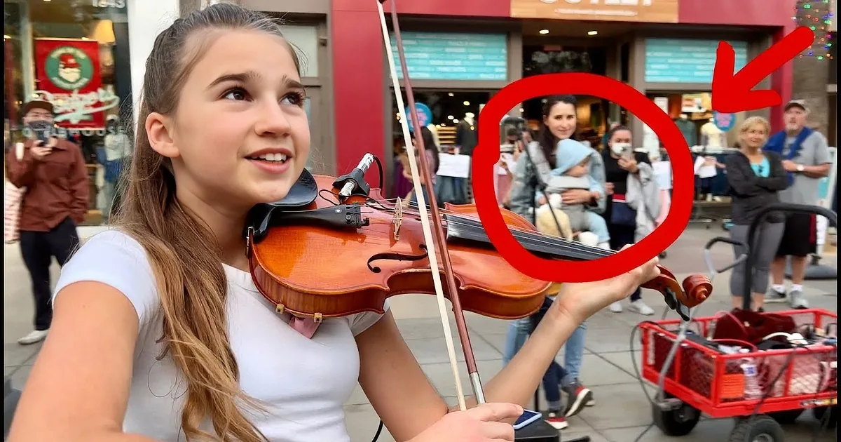 A young woman in a white shirt plays a violin in a bustling shopping area.