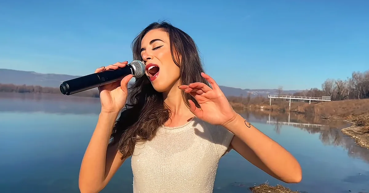 A woman sings passionately by a lake, holding a microphone, with a clear blue sky and distant hills in the background.
