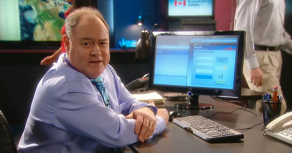 A man in a light blue shirt sits at a desk with a computer, looking at the camera.
