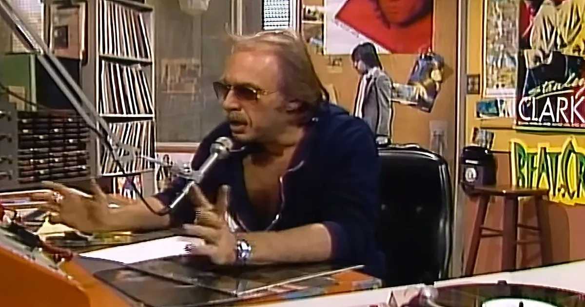 A man in sunglasses sits at a desk with a microphone, gesturing, in a room with vinyl records and posters.