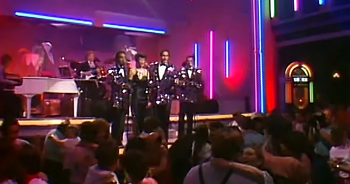 A band performs on stage with colorful neon lights and a lively crowd in the foreground.