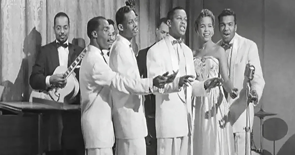 A group of men in white suits sing passionately, accompanied by a guitarist and a woman in a dress.
