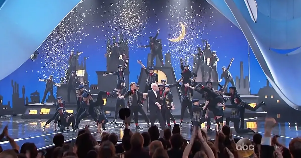 A man in a suit dances on stage with a group of performers, surrounded by confetti and a cityscape backdrop.