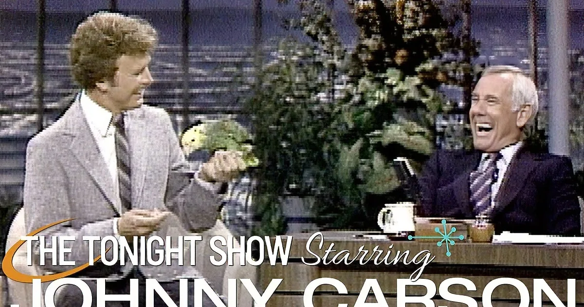 A man in a suit holds a parrot on his hand, smiling, while another man laughs on a talk show set.