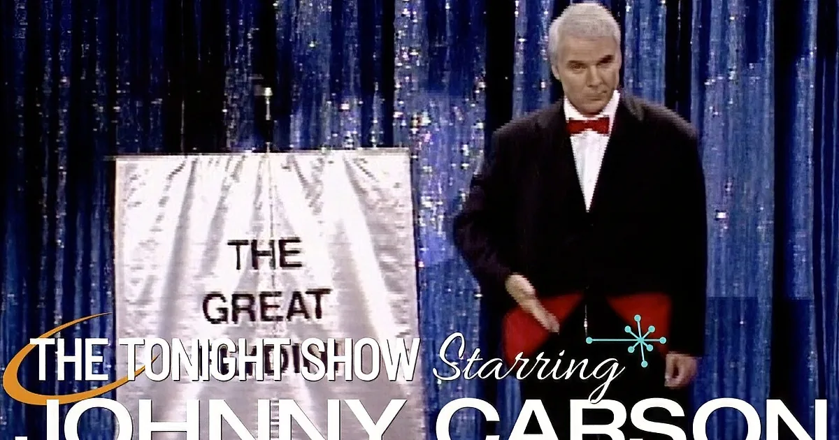 A man in a tuxedo and red bow tie raises his hand, standing against a blue curtain backdrop.