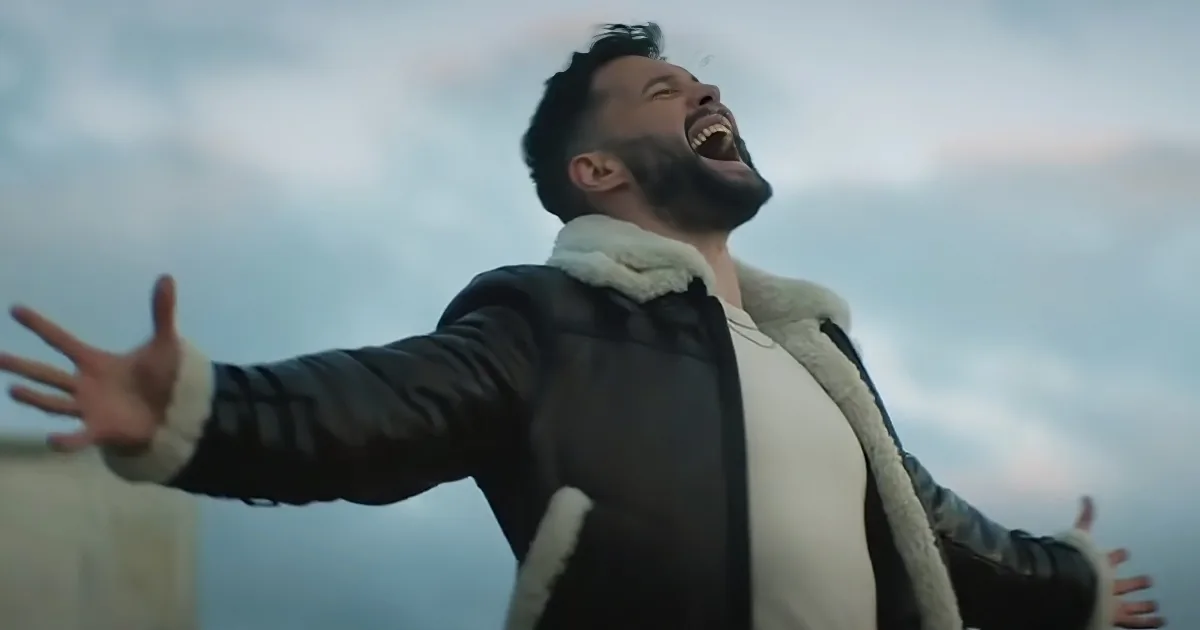 A man in a shearling-lined jacket with arms outstretched, laughing against a cloudy sky.