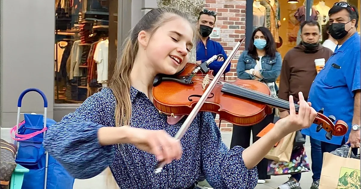 A young girl in a blue dress plays the violin on a sidewalk, surrounded by passersby and a store window.