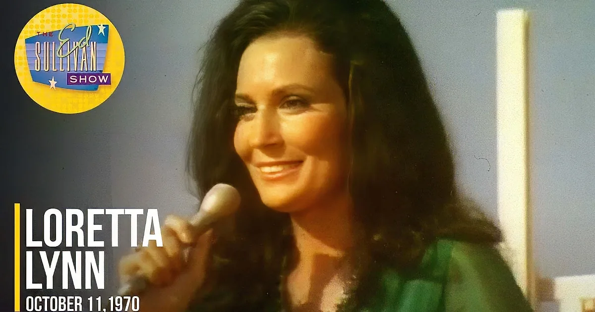 A woman with long dark hair sings into a microphone, wearing a green top, against a blue sky backdrop.
