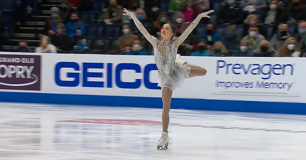 A figure skater in a silver outfit performs a pose on the ice with one leg extended.