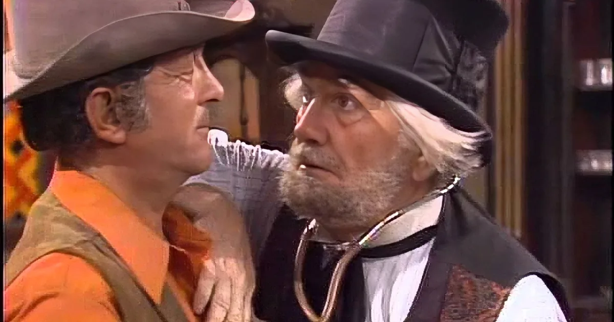 A man in a cowboy hat and vest laughs alongside another man in a vest and hat, holding a pipe.
