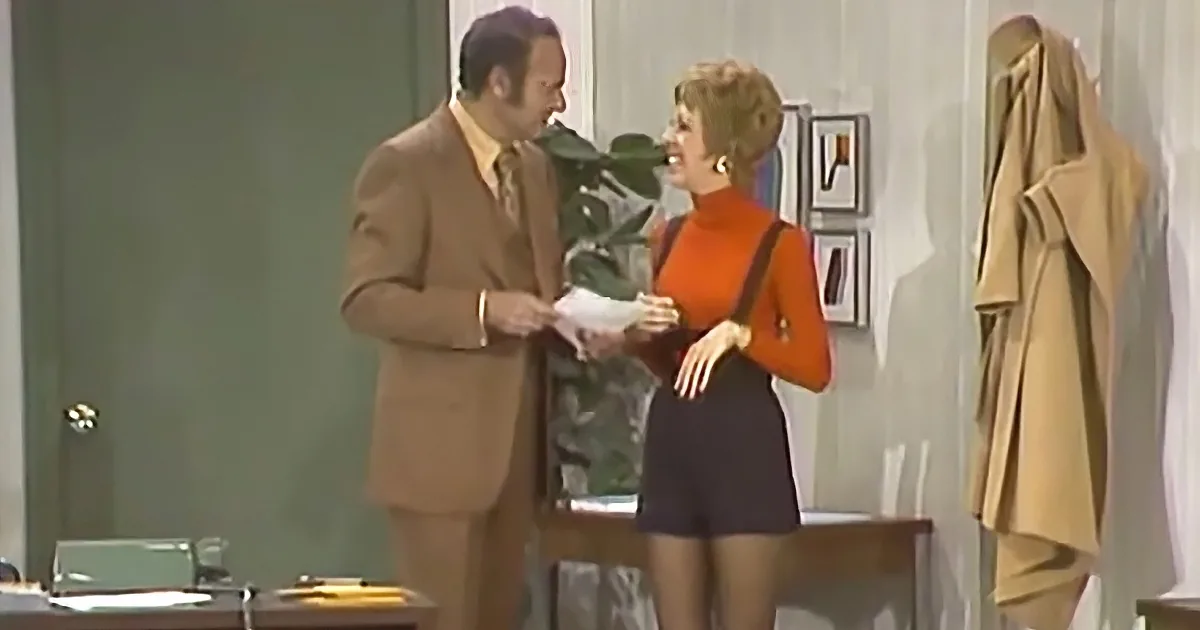 A man in a tan suit and a woman in an orange turtleneck are holding hands, standing in a room with a coat rack and framed...