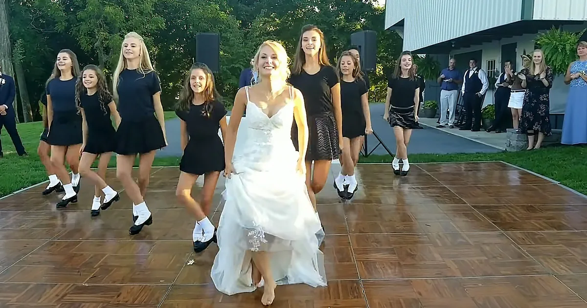 A bride dances on a wooden floor with a group of girls in black dresses and tap shoes.