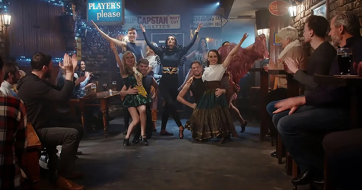 A group of dancers in costumes perform on a pub floor, surrounded by seated patrons.