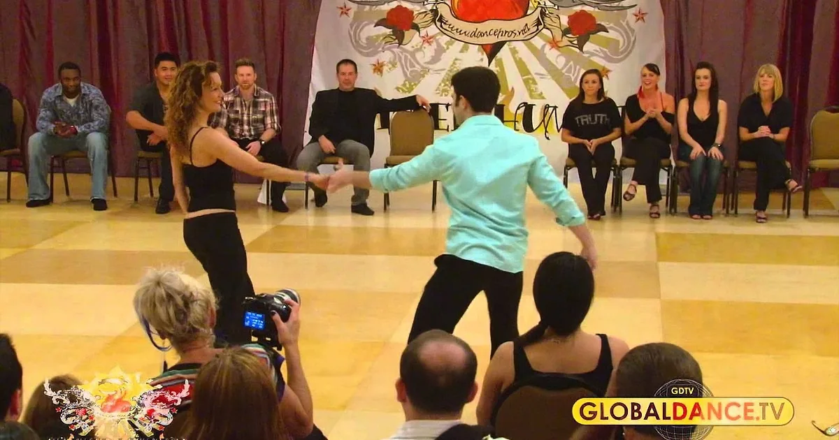 A man and woman are dancing, with the woman leading, in front of seated spectators.