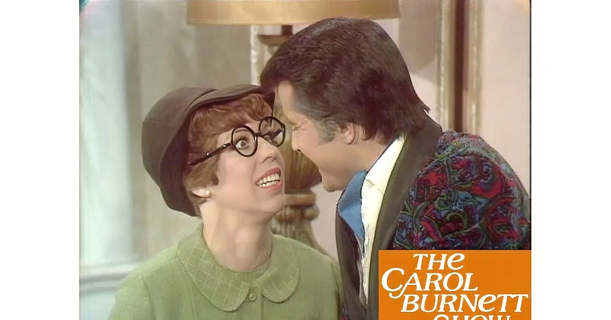 A woman in a green outfit and hat is speaking to a man in a floral suit, both appearing engaged in conversation.