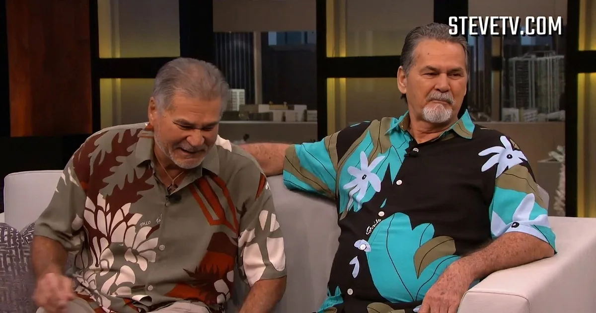 Two men in tropical shirts are seated on a white couch, one pointing and the other giving a thumbs-up.