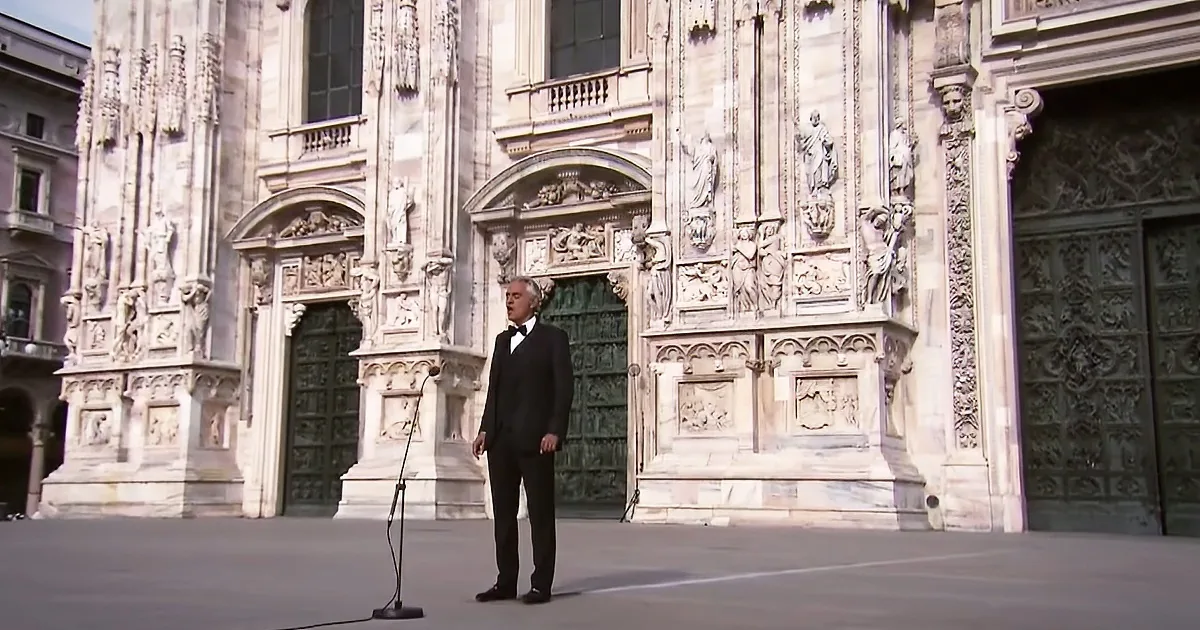 A man in a tuxedo stands in front of a grand, ornate cathedral, preparing to sing.