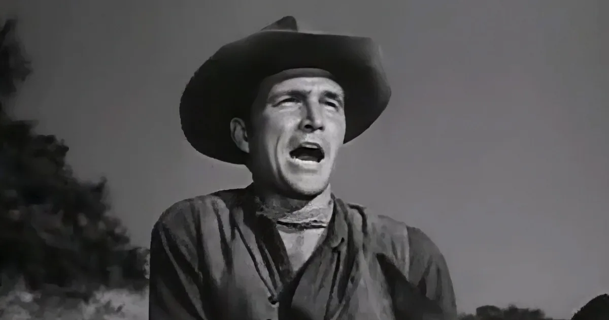 A cowboy in a wide-brimmed hat and shirt is shouting against a gray sky.