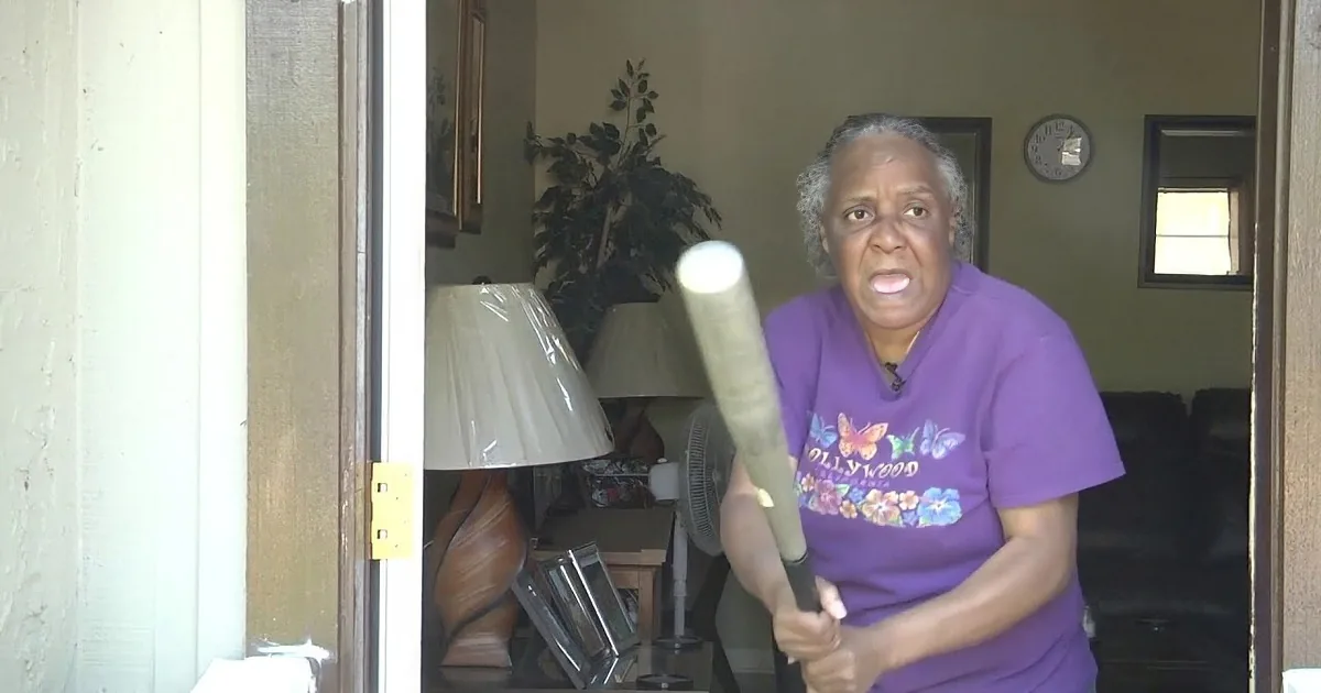 A woman in a purple shirt is holding a bat, appearing to swing it, with a lamp and a plant visible inside the house.