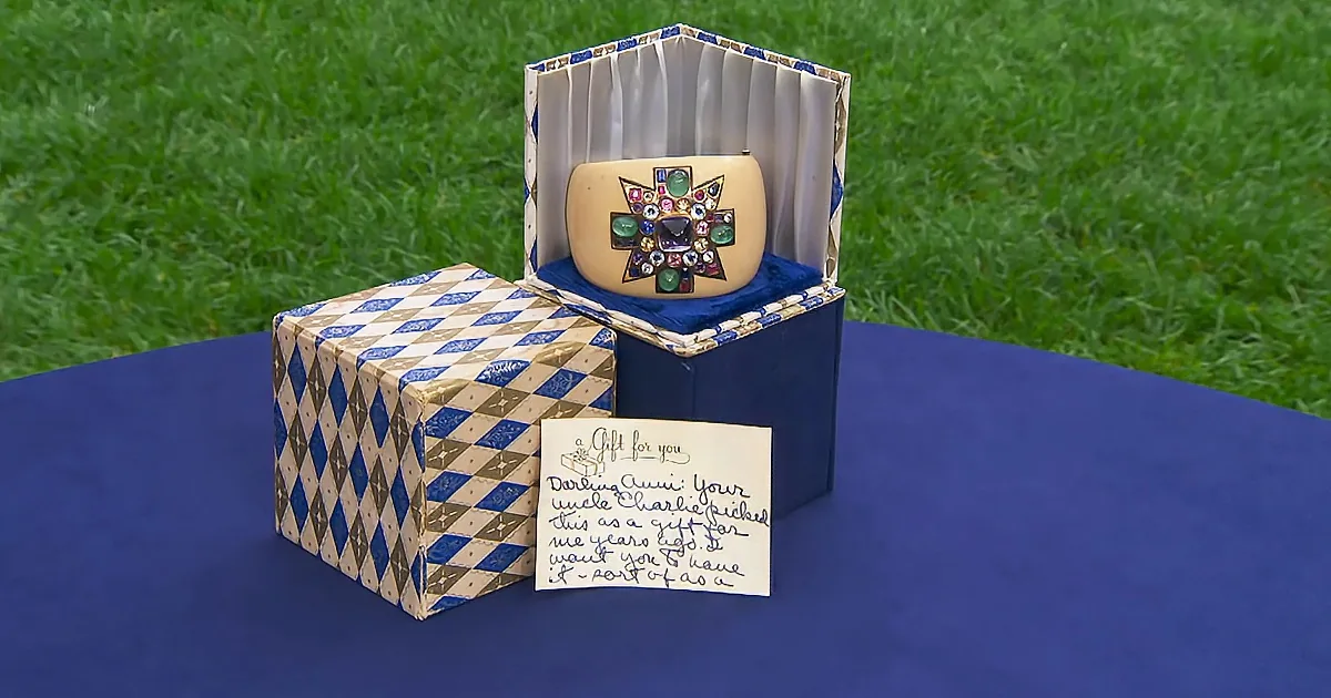 A decorative box with a note and a piece of jewelry inside is displayed on a blue table with grass in the background.
