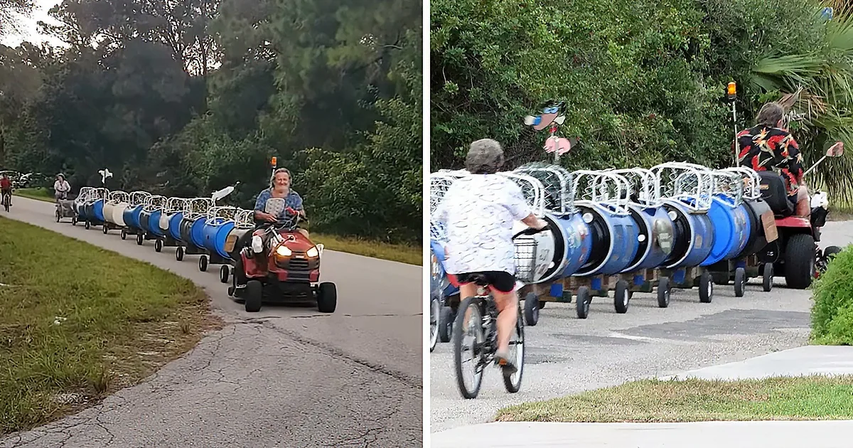 A person drives a red lawn mower pulling a trailer with blue barrels, followed by a cyclist.