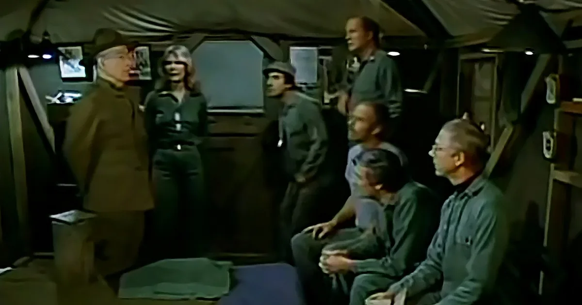 A group of people in a dimly lit room, some standing and some sitting, engaged in conversation.