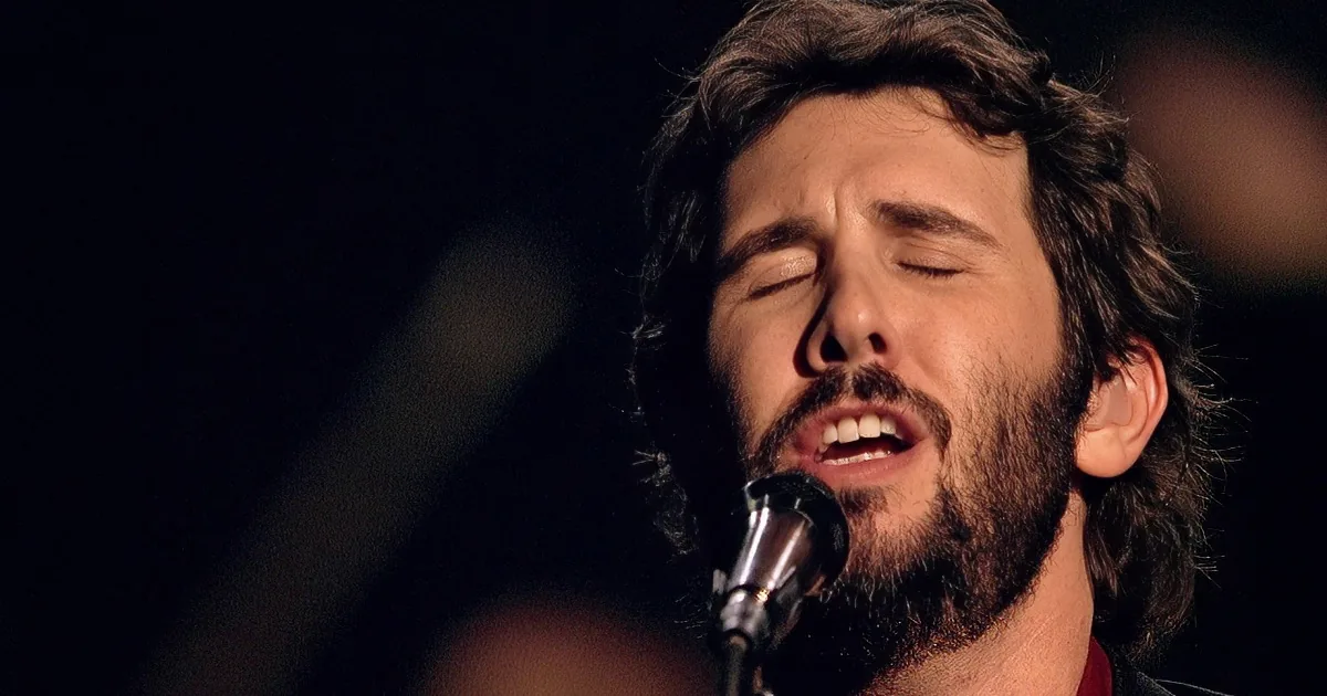 A man with curly hair and a beard sings passionately into a microphone, wearing a red shirt and black jacket.
