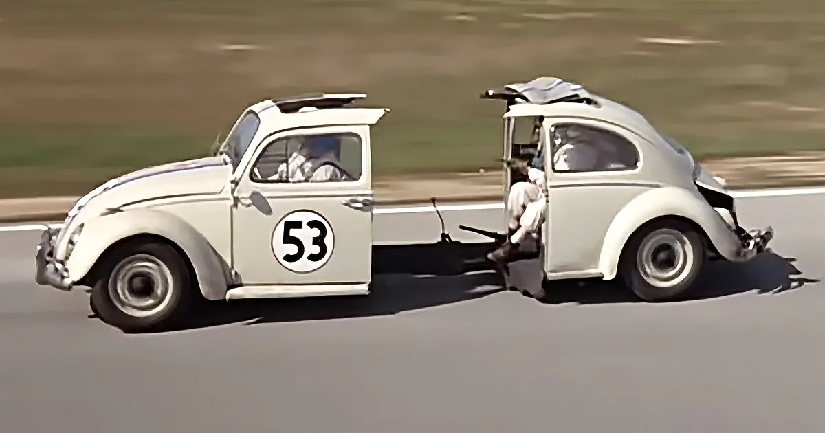 A white vintage car with number 53 on its door is towing another identical car on a road.