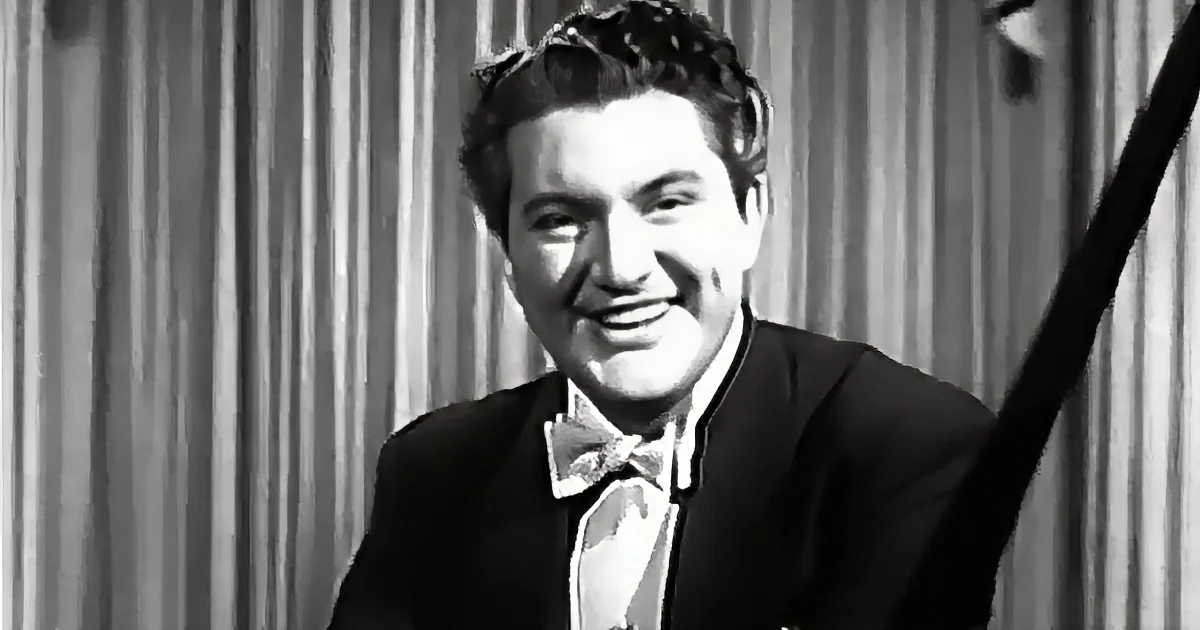 A man in a suit and bow tie smiles, holding a microphone, against a curtain backdrop.