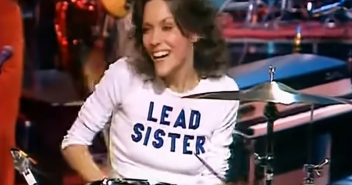 A person wearing a "LEAD SISTER" shirt smiles while playing drums in a band setting.