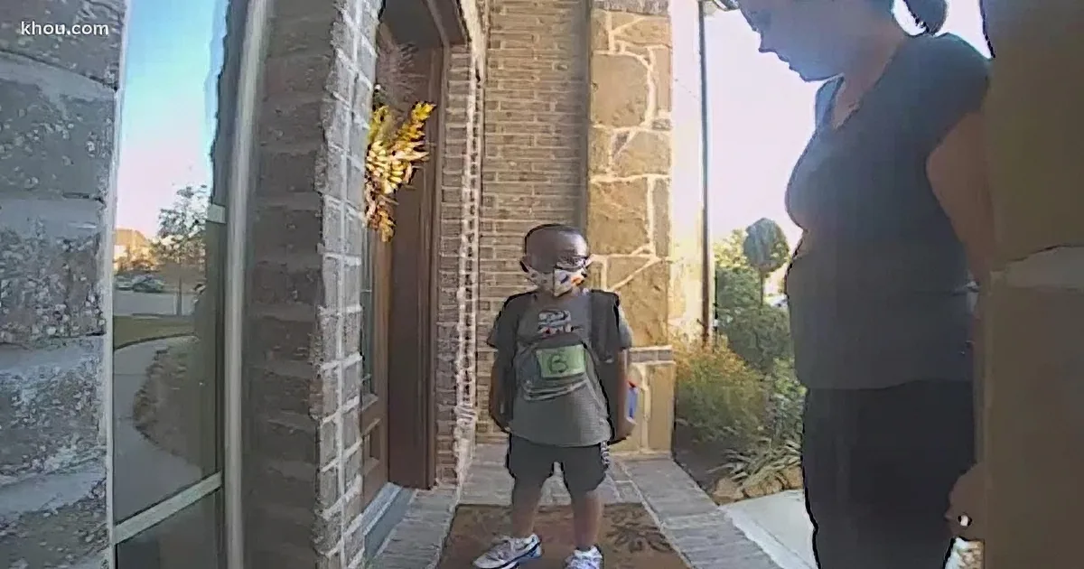 A young boy wearing a mask and backpack stands in front of a house entrance, with an adult nearby.