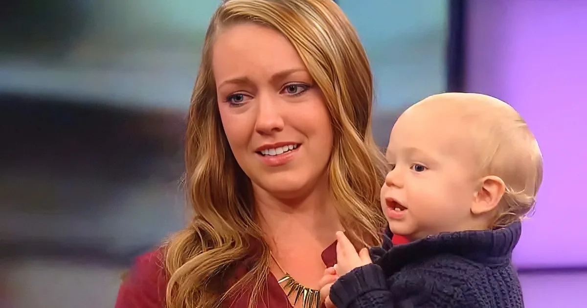 A woman with long blonde hair holds a baby in a dark sweater, both smiling.