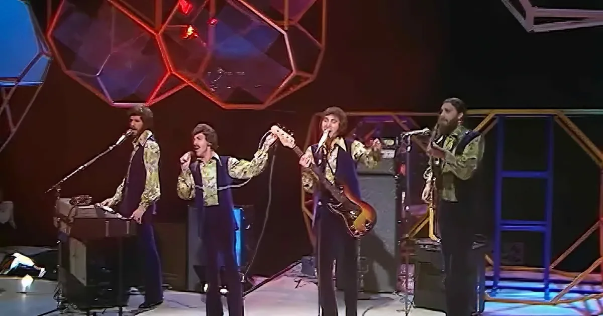 A band performs on stage with colorful geometric lights and microphones.