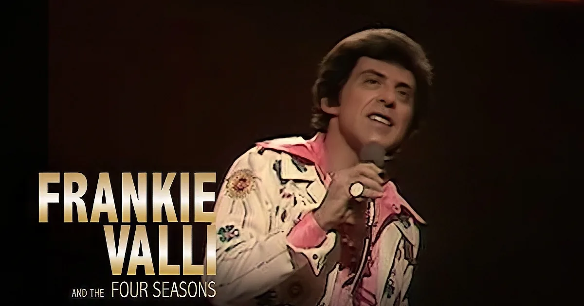A person in a floral pink and white shirt holds a microphone on stage.