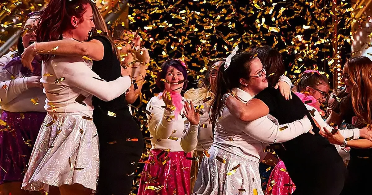 Two women in white and silver dresses hug amidst golden confetti, surrounded by a festive crowd.