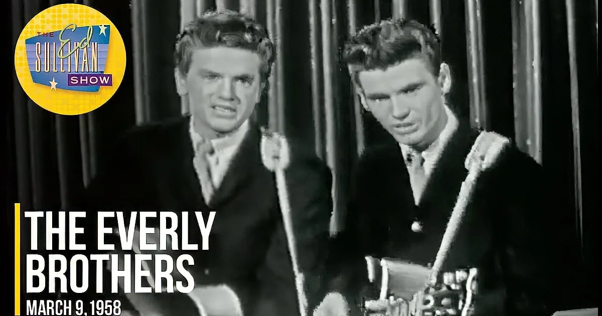 Two men in suits play guitars, one with a serious expression, against a dark, vertical-striped background.