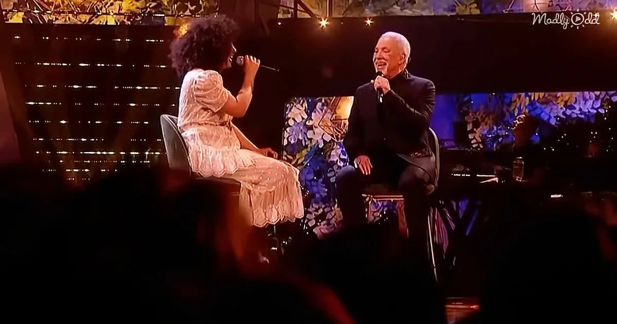 A woman in a white dress and a man in a black suit are seated on stage, each holding a microphone.