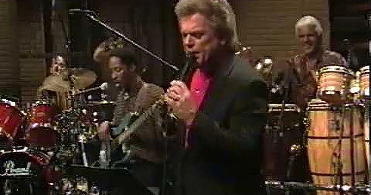A man in a suit sings passionately on stage with a band, including a drummer and guitarist.