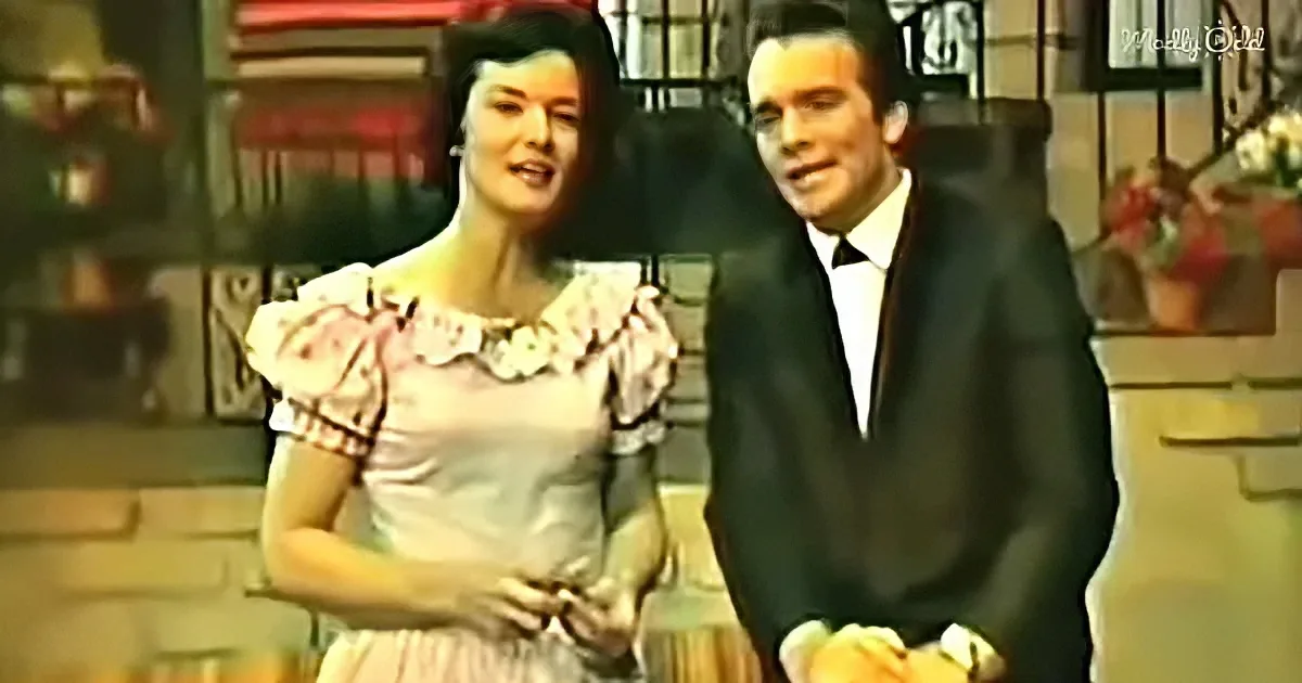 A woman in a pink dress and a man in a suit stand together, smiling, with a decorative background.