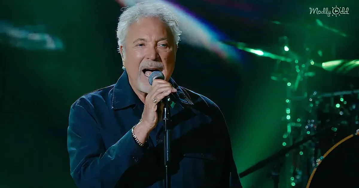 A man in a dark jacket sings into a microphone on stage with green lighting.