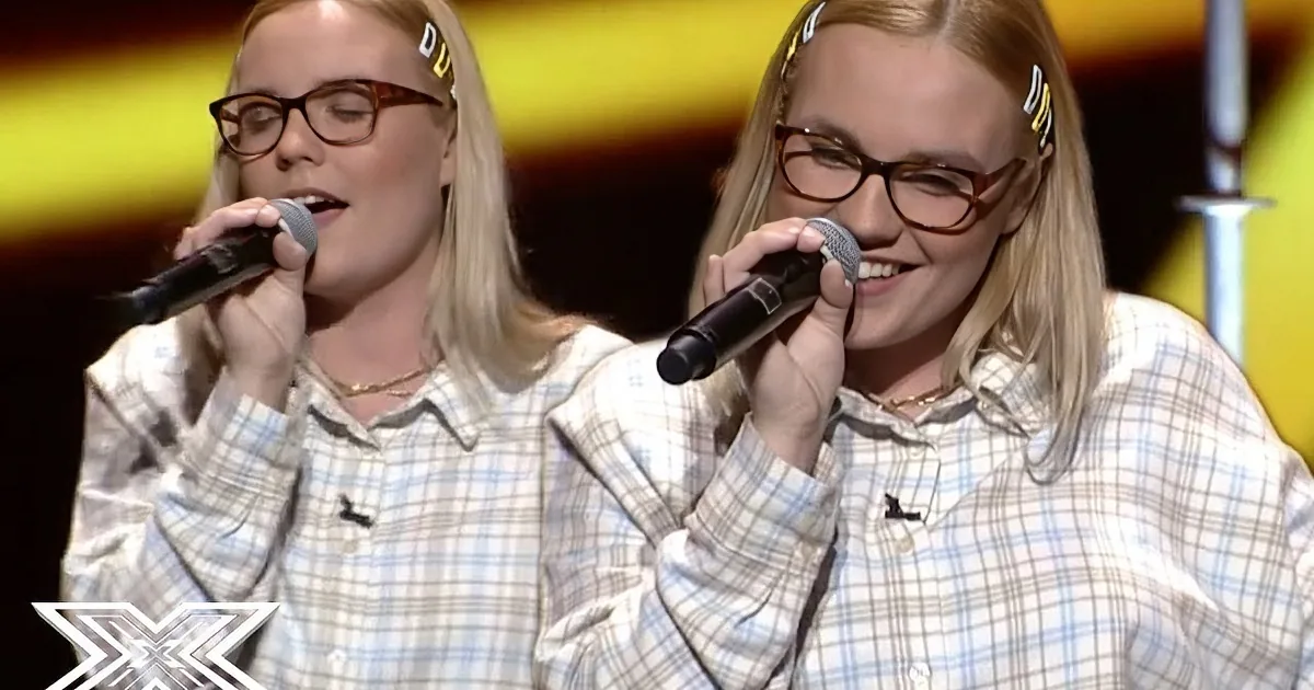 A woman with glasses sings into a microphone, wearing a plaid shirt, on a stage with a colorful background.