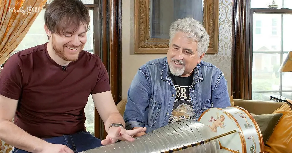 Two men smile, one in a maroon shirt, the other in a denim shirt, interacting with a large, ornate object.