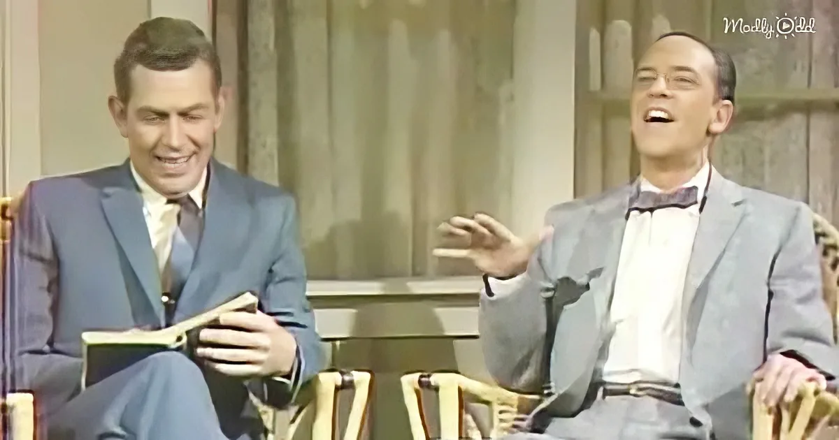 Two men in suits, one reading, the other gesturing, seated in a studio setting.