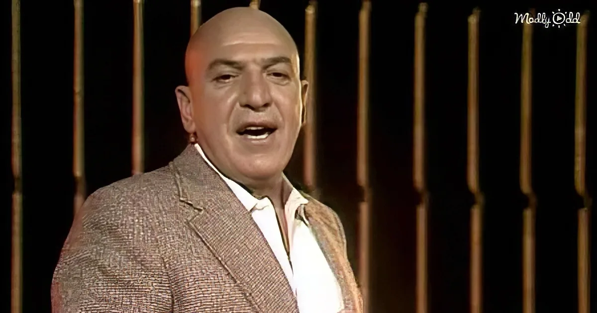 A man in a suit and white shirt stands against a dark background with vertical golden bars.