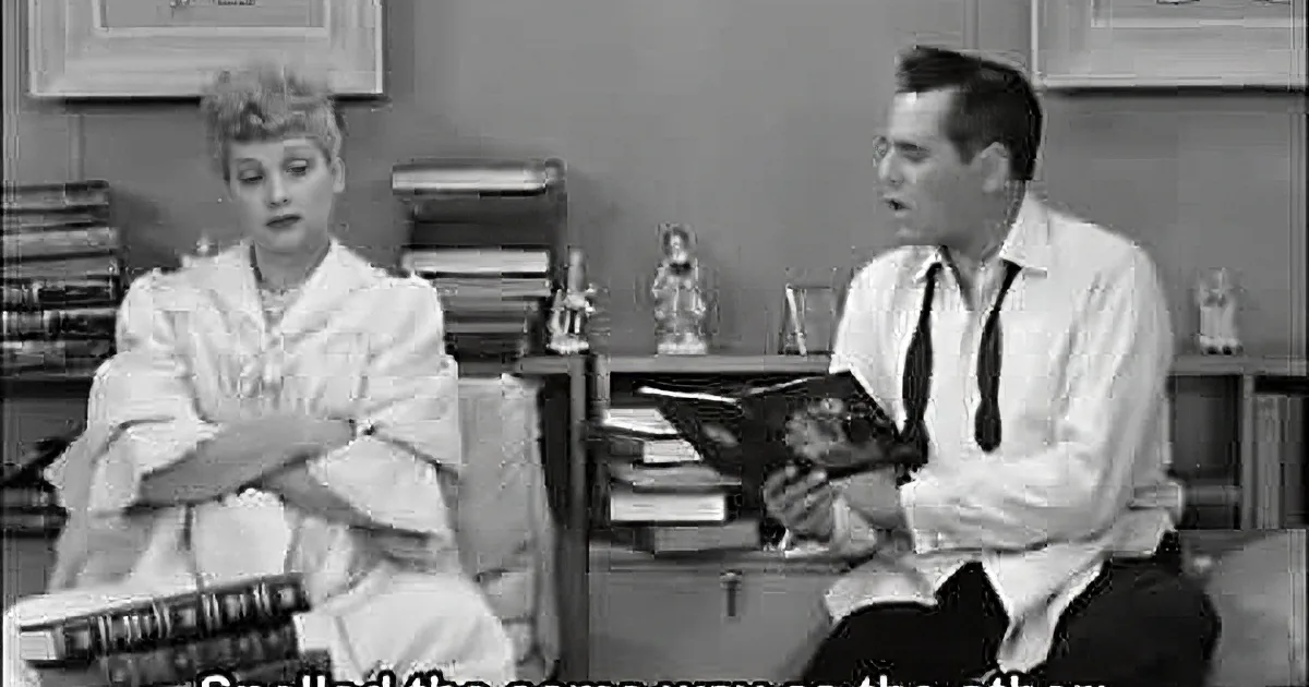 Lucille Ball sits with arms crossed, smiling, while Desi Arnaz holds a clipboard, both in a vintage office setting.