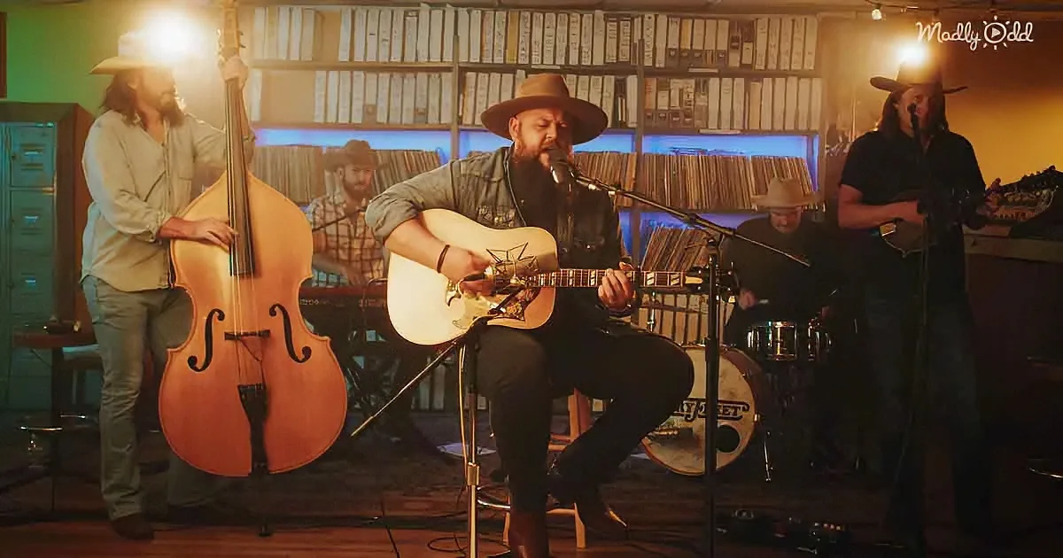 A man in a hat plays guitar in front of a band in a dimly lit room with vinyl records.