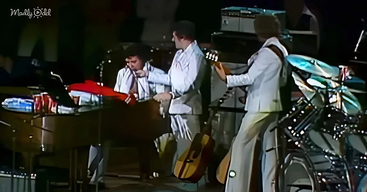 A musician in a white suit plays guitar on stage with other musicians and instruments.