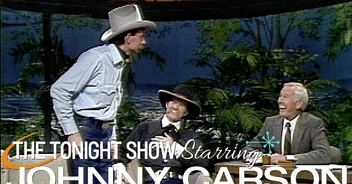 A man in a cowboy outfit and hat is gesturing with his hands, while another man in a suit smiles in the background.