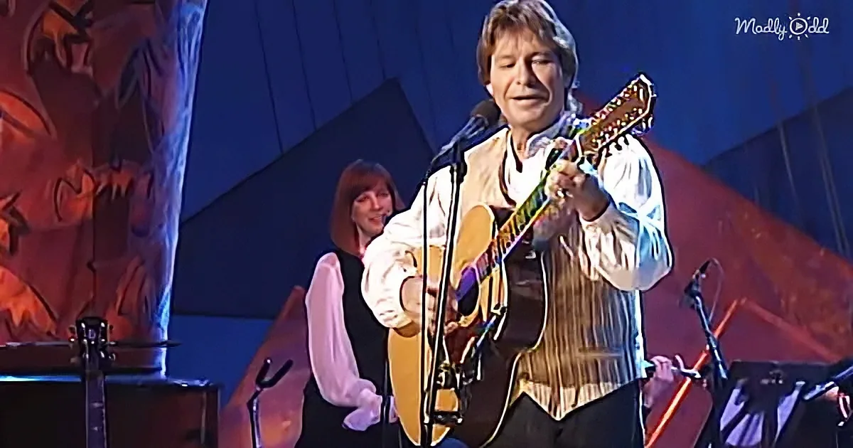 A man plays guitar on stage, singing into a microphone, with a woman in the background.