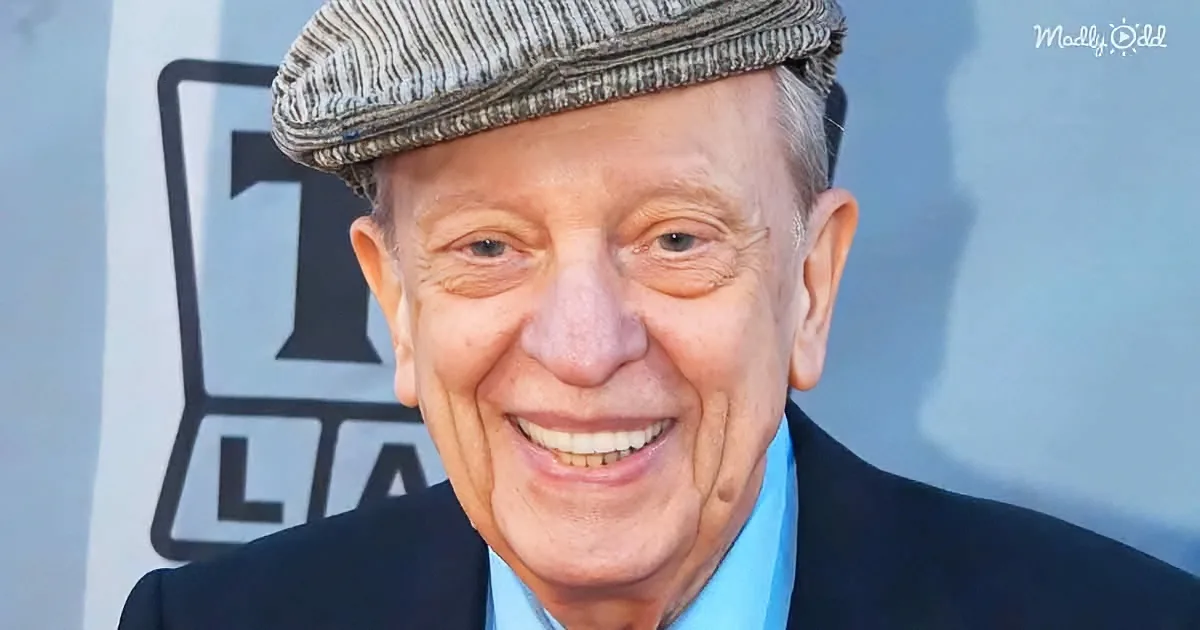 A smiling older man in a suit, flat cap, and blue shirt, with a blurred background.